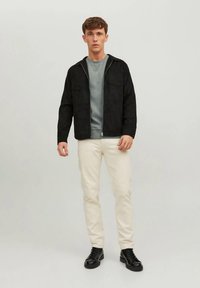 Melna jaka ar rāvējslēdzēju virs pelēka T-krekla, kopā ar krēmkrāsas taisna piegriezuma biksēm un melnām kurpēm ar šņorēm. Vienkāršs fons.