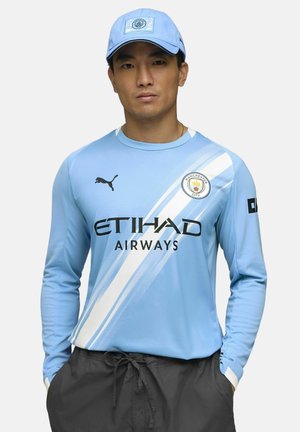 Lyseblå langærmet trøje med hvide skråstriber, der har "ETIHAD AIRWAYS," Manchester City-logoet og sort Puma-logo. Kombineret med en matchende kasket.