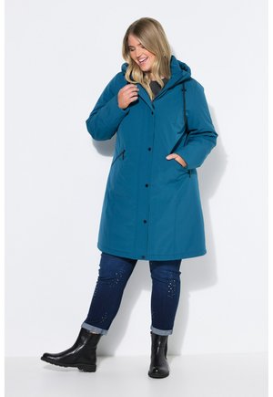 Femme souriante portant un manteau à capuche bleu sarcelle, un jean foncé avec des ornements et des bottines noires, debout devant un fond uni.