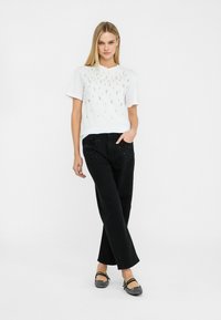 T-shirt bianco a maniche corte con dettagli decorativi di perline, abbinato a pantaloni neri con decorazioni vicino alle tasche e scarpe nere flat.