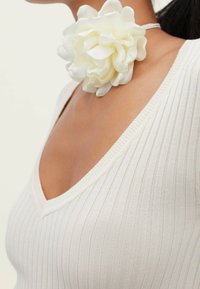 Vrouw in een witte geribbelde V-hals top en een grote witte stoffen bloem als choker ketting tegen een effen achtergrond.