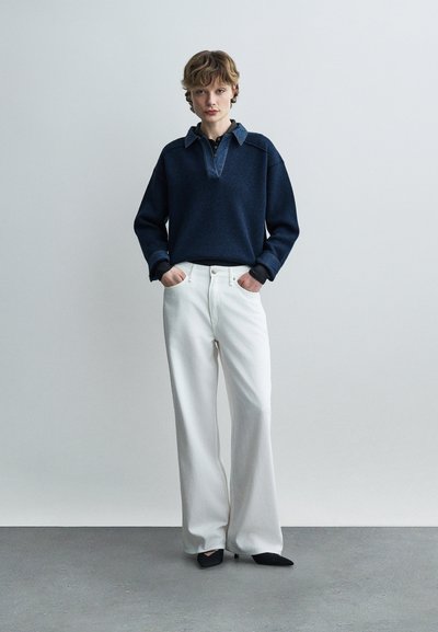 Personne debout les mains dans les poches portant un pull à col bleu marine, un pantalon large blanc et des chaussures pointues noires sur un fond uni.