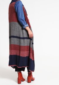 En person som bär en lång, randig stickad väst i röd, grå och marinblå över en blå skjorta med uppvikta ärmar, svarta byxor och röda klackstövlar.