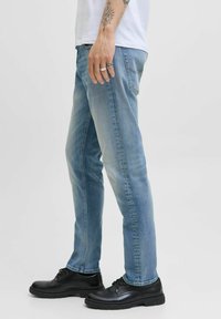 Hombre que lleva puesta una camiseta blanca, jeans de mezclilla azul claros y zapatos negros de cordones, con tatuajes en la mano y el antebrazo, así como anillos visibles.
