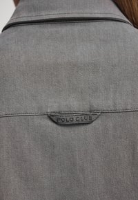 Grijze denimjack met een kraag en een opgenaaid label met de tekst "POLO CLUB." De stof heeft een gladde textuur en een gestructureerde stiksel.