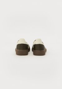 Sneakers z kremową cholewką i ciemnozielonymi akcentami, teksturowaną brązową podeszwą i prostym kształtem. Widok z tyłu podkreśla kontrast kolorystyczny.