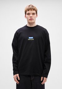 Μαύρη μακρυμάνικη μπλούζα με στρογγυλή λαιμόκοψη. Διαθέτει κεντρικό λογότυπο: "HUGO" σε μπλε και "JOHN K." σε χρυσό. Απλός σχεδιασμός, από βαμβακερό υλικό.