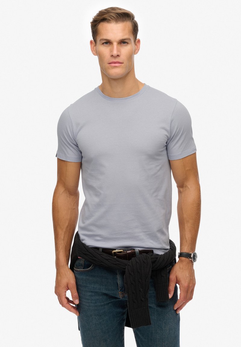 Lysegrå t-shirt med korte ærmer, tætsiddende design, båret med en mørk strikket sweater bundet om livet, kombineret med blå jeans og et ur.