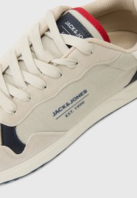 Beige sneaker with canvas and suede materials, featuring navy accents, round laces, and branding details in blue on the side. Red collar trim.