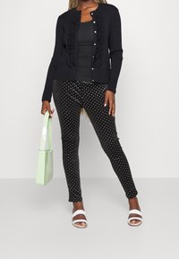 Femme portant un gilet noir à volants et un pantalon à pois, tenant un sac fourre-tout vert clair, debout avec une jambe légèrement pliée.