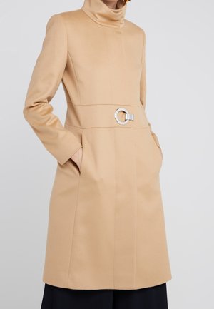 Vrouw die een beige knie-lang mantel draagt met een hoge kraag, zijzakken en een zilverkleurige ronde gesp aan de taille.