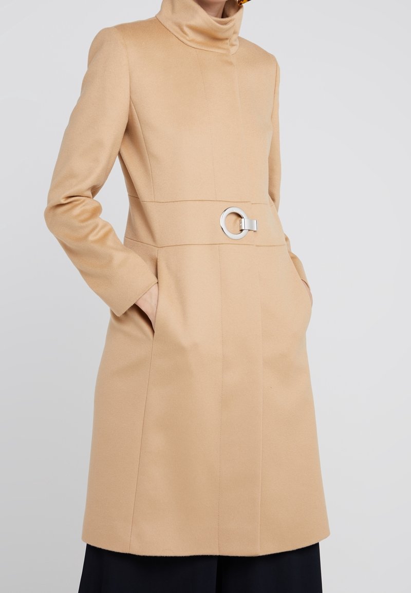 Femme portant un manteau beige longueur genou avec un col montant, des poches latérales et une ceinture à boucle circulaire argentée à la taille.