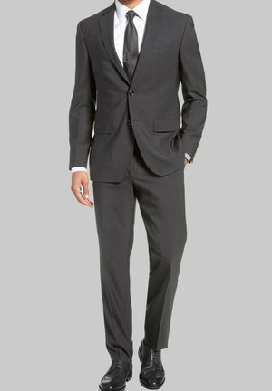 Costume gris avec une veste à deux boutons, un pantalon à plat et une chemise habillée blanche. Porté avec une cravate noir texturée et des chaussures de ville noires.