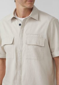 Beige korte mouw shirt van lichte stof met een puntkraag, sluiting met knopen aan de voorkant en twee borstzakken. Bevat marineblauwe knoopaccenten.
