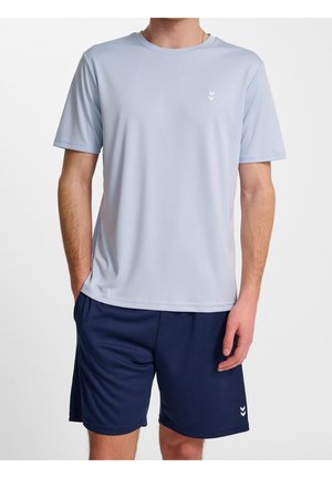 Mann trägt hellblaues kurzärmeliges Hemd und dunkelblaue Shorts, steht mit einer Hand in der Tasche vor weißem Hintergrund.