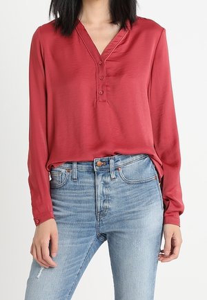 Woman wearing a long-sleeve red buttoned blouse tucked into light blue high-waisted jeans with slight distressing.