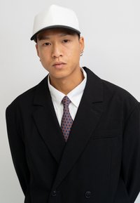 Black double-breasted blazer over a white shirt, featuring a patterned tie with circles. A white cap completes the look.