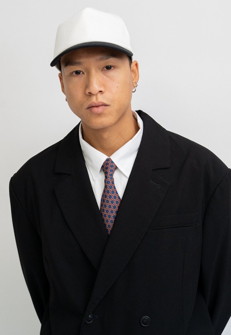 Black double-breasted blazer over a white shirt, featuring a patterned tie with circles. A white cap completes the look.