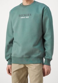 Man i en teal sweatshirt med texten "DICKIES SINCE 1922" på bröstet och beige byxor, stående mot en enkel bakgrund.