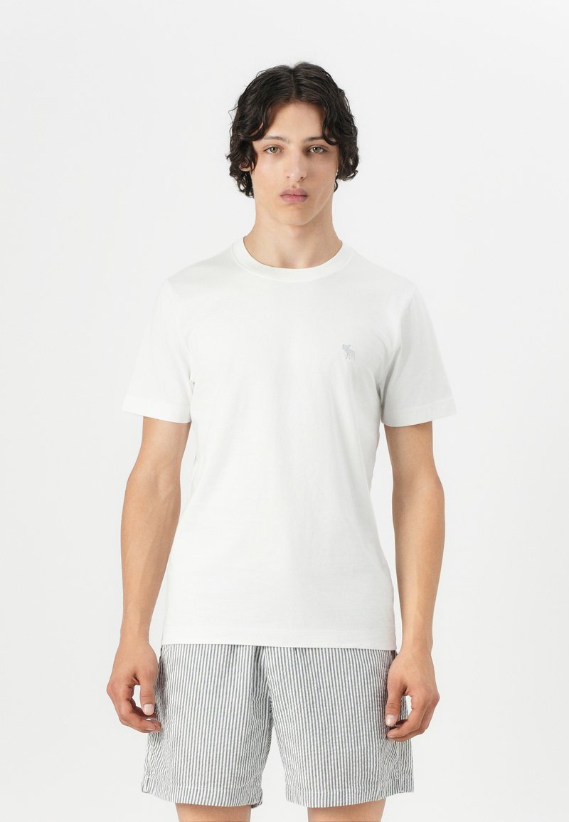 Hvid bomulds-T-shirt med korte ærmer, rund halsudskæring, med et lille gråt logo. Matchet med grå- og hvide stribede shorts.