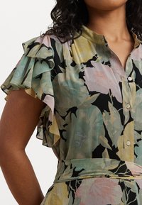 Blomsterbluse med volangerm, utstyrt med pastellfarger på en svart bakgrunn, knappestenging og belted midje. Strukturert stoff.