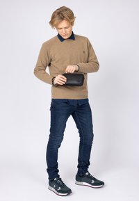 Maglione a maglia marrone sopra una camicia blu con colletto, jeans slim-fit blu e sneakers grigie. Tiene una piccola borsa nera strutturata con un gancio.