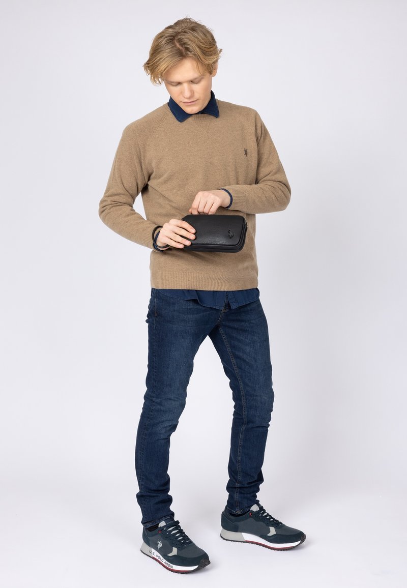 Maglione a maglia marrone sopra una camicia blu con colletto, jeans slim-fit blu e sneakers grigie. Tiene una piccola borsa nera strutturata con un gancio.