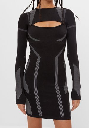 Vrouw draagt een nauwsluitende zwarte jurk met lange mouwen, geometrische witte streeppatronen en een uitsnijding op de borst.