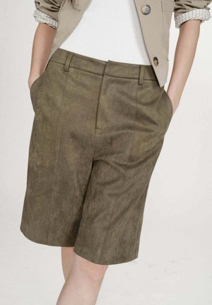 Femme portant un short en suède vert olive mi-longueur avec des poches latérales, associé à une veste beige et un haut blanc côtelé, les mains dans les poches.