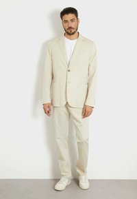 Blazer de color beige claro y pantalones a juego, hechos de algodón, que cuentan con un cierre de un botón, bolsillos de parche y un ajuste relajado. Zapatillas blancas casuales.