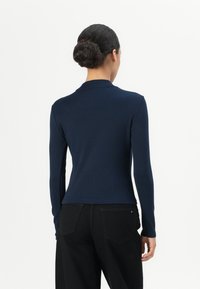 Maglietta a maniche lunghe blu navy realizzata in tessuto morbido, con collo a camicia e design aderente. Abbinata a jeans neri, che mostrano la vista posteriore.