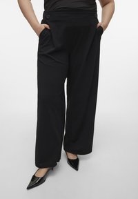 Pantalons larges noirs en tissu lisse. Dotés de deux poches latérales et d'accents de boutons sur la ceinture. Associés à des escarpins noirs à bout pointu.