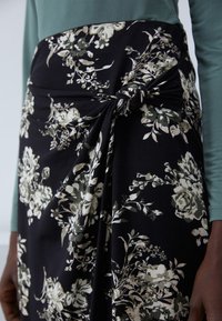 Jupe portefeuille noire à motifs floraux avec un détail en nœud, mettant en avant un mélange de fleurs blanches et vertes. Tissu doux avec une texture lisse.
