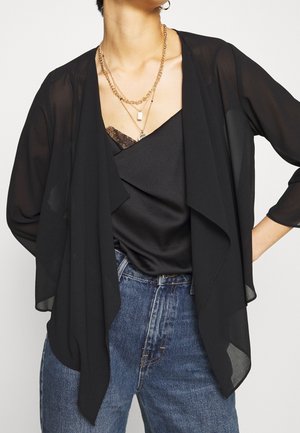 Sort gennemsigtig bolero med draperet front, lange ærmer, parret med en sort satin-top og jeans, med lagdelte guldhalskæder.