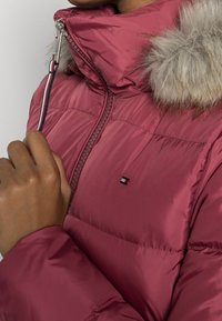 Chaqueta acolchada burdeos con capucha de pelo sintético. Presenta un cierre delantero con cremallera y un parche con logotipo. La tela tiene una textura suave y un acabado brillante.
