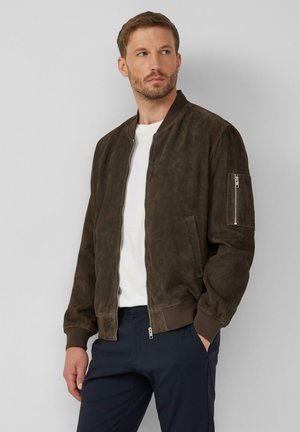 Braune Wildleder-Bomberjacke mit gerippten Bündchen und Saum, Reißverschlusstasche am Ärmel und zwei Seitentaschen. Kombiniert mit einem weißen T-Shirt und dunklen Hosen.