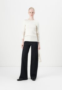 Helles beige Langarmoberteil mit gerippter Textur, kombiniert mit dunkelblauen Weitenbeinen-Jeans und einer kleinen weißen Handtasche. Schwarze Ballerinas.