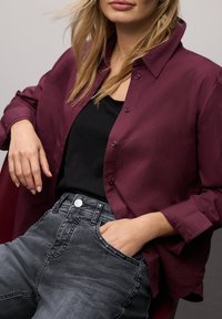 Bordowa koszula na guziki nałożona na czarny top, zestawiona z wysokim stanem ciemnoszarymi jeansami z ozdobnymi guzikami.