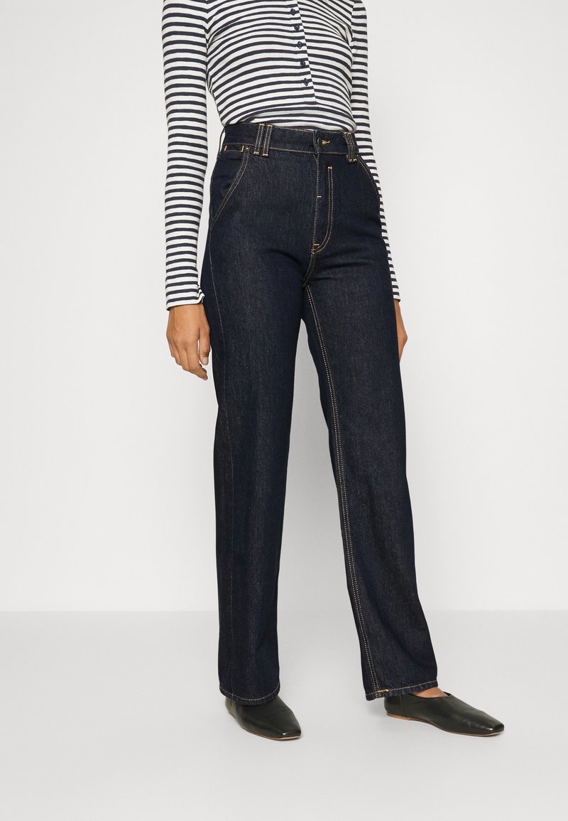 Femme portant un jean droit taille haute foncé, des ballerines noires pointues et un haut blanc à manches longues avec des rayures horizontales noires.