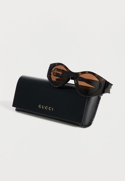 Γυαλιά ηλίου στυλ γατίσια μάτια με σχέδιο χελώνας και καφέ φακούς, τοποθετημένα πάνω σε μαύρη θήκη Gucci με χρυσό λογότυπο σε λευκό φόντο.