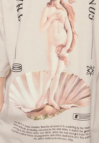 Camisa color beige claro con una impresión de "El nacimiento de Venus" sobre un fondo de conchas marinas, colores delicados y texto impreso sobre la obra de arte.