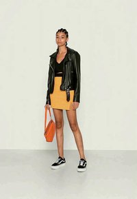 Jaqueta de motoqueiro de couro preto, saia mini texturizada amarela, bolsa laranja e ténis pretos. Modelo está encostada a um fundo claro.