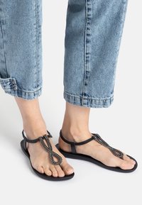 Black sandals with a flat sole, featuring a chain-like decorative strap and a simple T-bar design. Smooth texture with adjustable ankle straps.