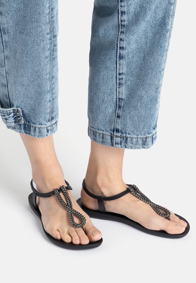 Black sandals with a flat sole, featuring a chain-like decorative strap and a simple T-bar design. Smooth texture with adjustable ankle straps.