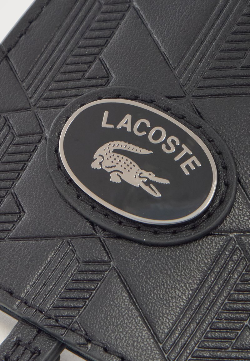 Primo piano di un oggetto nero testurizzato con una targhetta in metallo con il logo Lacoste raffigurante un alligatore e motivi geometrici cuciti.