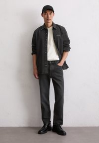 Man in een zwarte baseballcap, een zwart denimshirt over een wit T-shirt, zwarte jeans en zwarte leren schoenen, staande tegen een witte textuur muur.