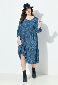 Sininen kukkakuosiset mekko, jossa on kootut vyötäröt, pitkät hihat ja sivuaukko; yhdistettynä mustiin nauhakenkiin ja tummaan hattuun.