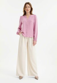 Hellrosa Hemdbluse mit langen Puffärmeln und Stickerei, kombiniert mit weißen weit geschnittenen Hosen und beigen spitzen Schuhen.