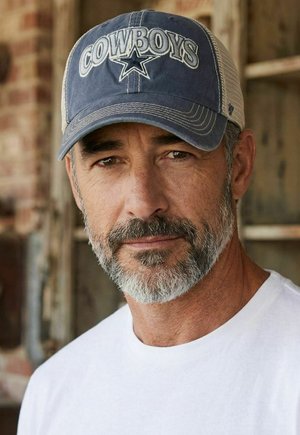 Middle-aged man with salt-and-pepper beard wearing a blue Cowboys cap and white shirt, standing by a rustic wooden wall.