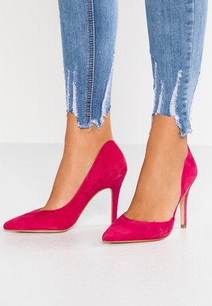 Fødder iført lyserøde semskede stiletter parret med lyseblå, slidte ankellange jeans mod en ensfarvet baggrund.